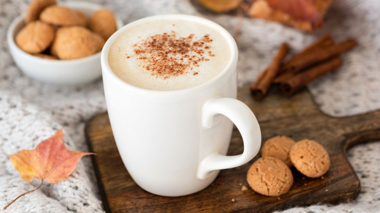 A pumpkin spice latte in a white coffee mug on a board next to biscotti and autumn leaves