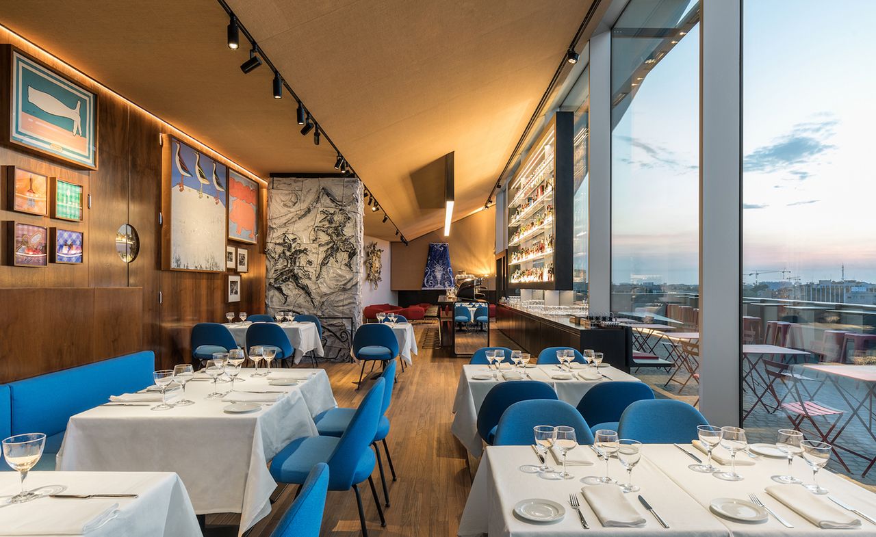 View of the interior of the restaurant with tables covered in a white tablecloth, blue chairs, and wood details on the floor and walls, with a view over the skyline of the city.