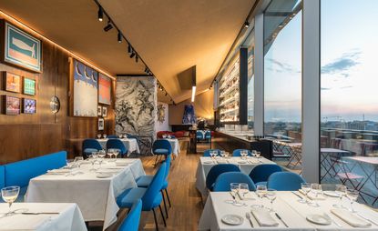 View of the interior of the restaurant with tables covered in a white tablecloth, blue chairs, and wood details on the floor and walls, with a view over the skyline of the city.