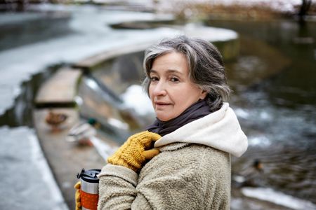 An older woman is bundled up outside with a hot drink, turning back toward the camera.