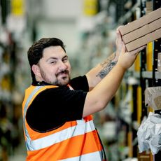 A man working in a warehouse lifts a stack of cardboard boxes from a shelf. He wears an orange high-visibility vest over a black shirt, has short dark hair, a beard, and a visible tattoo on his right arm.