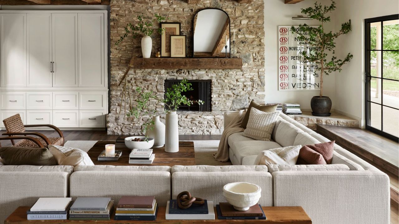 A white sectional sofa, covered with white and beige throw pillows, encircling a wooden coffee table; in the background, a fireplace in a stone wall; to the left, white cabinetry; to the right, French doors opening onto a garden view. 