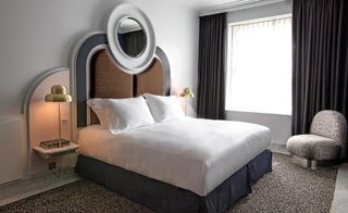 A room in the Henrietta Hotel. A large bed with white linen and a rounded headboard in deep red and black is to the left, next to which is a night table with a golden lamp. To the far wall, there is a window that lets in a lot of light.