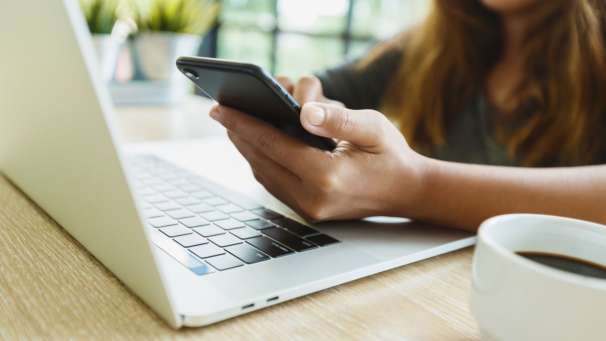 A person holding an iPhone next to an Apple computer, representing an article about how to transfer photos from an iPhone to a computer