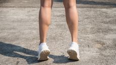 Person's bare legs below the knees seen from behind. They stand on tip toes and are wearing white trainers