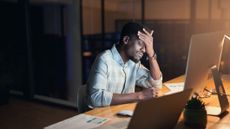 A stressed looking man sat at a desk with his head in his hand, sat in front of a monitor late at night.