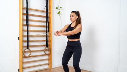 A woman performs the Pallof press in a white studio sport. She stands with her knees slightly bend holding a resistance band attached to wooden wall bars in front of her chest. She wears her hair in a pony tail and is wearing black sports leggins and a cropped black tank top.