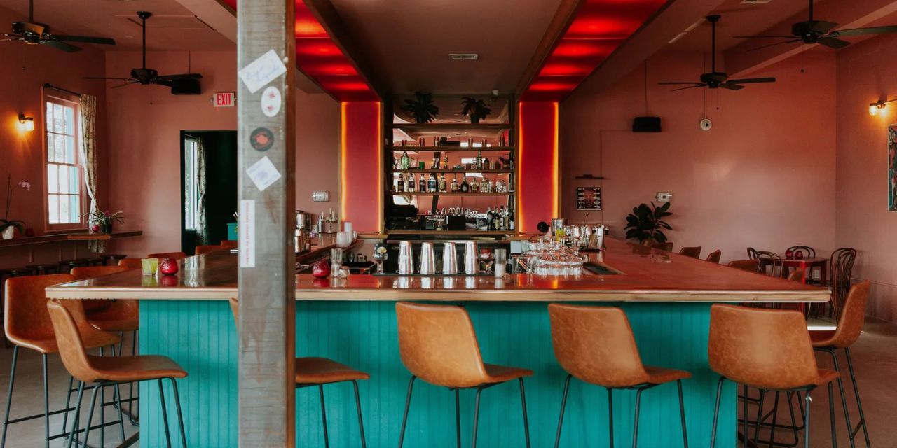 The bar counter of a New Orleans cocktail bar features sea green wood paneling, mid-century modern leather stools, a terracotta background, and a lit-up, softly glowing red wall accent and ceiling.