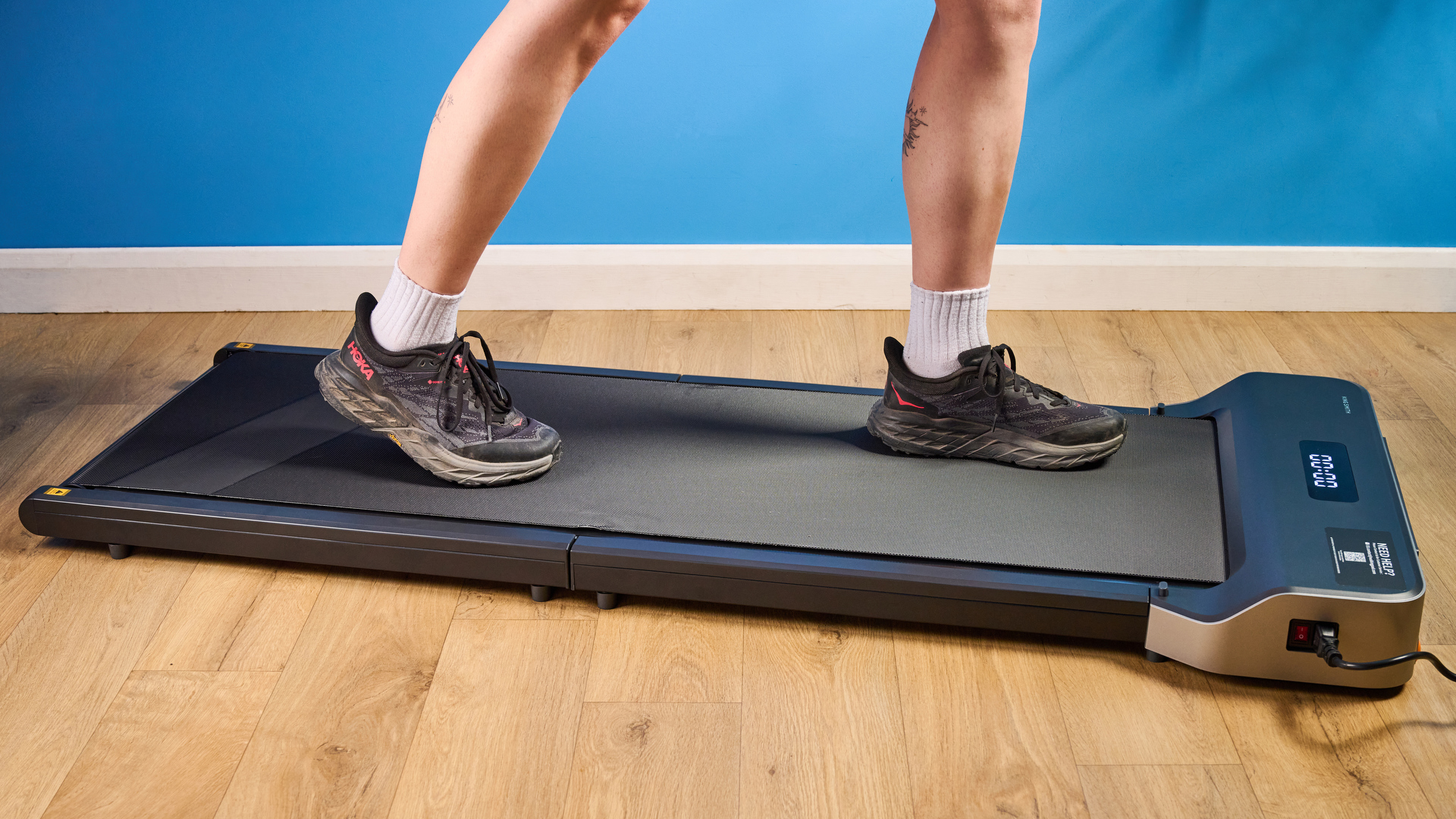 A woman walking on the WalkingPad Z1 wearing Hoka running shoes photographed in front of a blue background. 