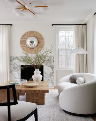 White living room with marble fireplace, burled wood coffee table, curved white boucle sofa and round wood mirror