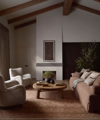 Living room with high ceiling, wood beams, dusty pink sofa, boucle white armchairs, low wood coffee table and dark orange patterned rug