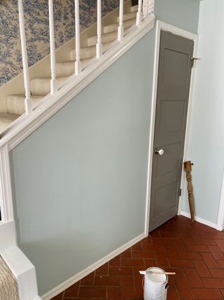 a plain entryway, with red/orange tiled floor, a plain blue wall, and beige stairs, with floral blue and white wallpaper. A pot of paint also sits on the floor
