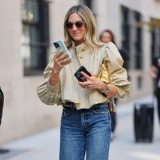 NYFW street style photo of a woman wearing a puff-sleeve khaki jacket and jeans while walking and looking at her phone
