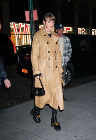 Taylor Swift wearing a tan leather trench coat with black heeled booties and a black shoulder bag