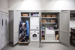 grey and white utility room with large cupboards with with washing machines inside by Burlanes, and baskets of storage, and a clothes dryer