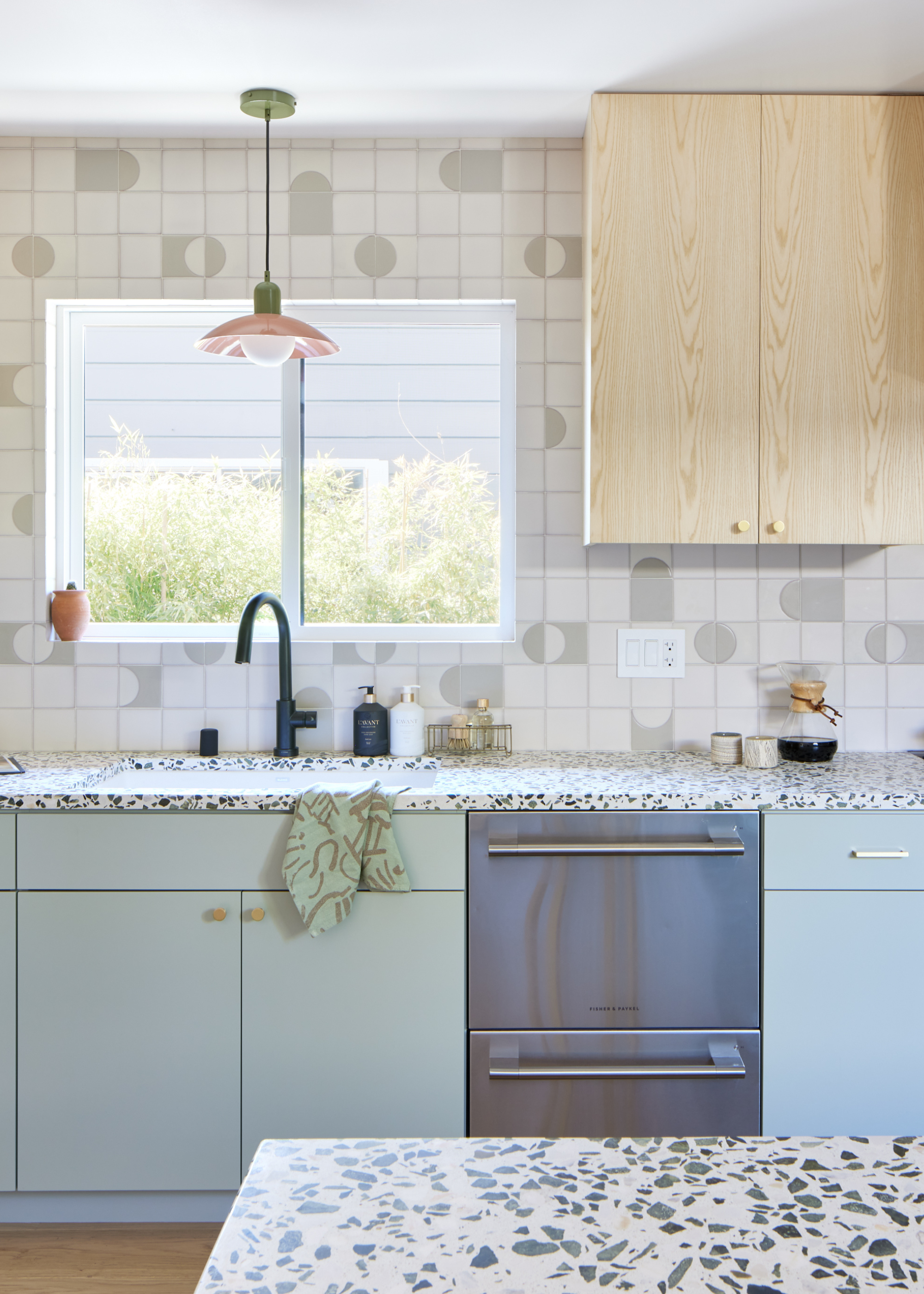 A bright kitchen with a tile backsplash, warm wood cabinetry, a hanging pendant light, terrazzo counters, sage green cabinetry, and a window by the sink