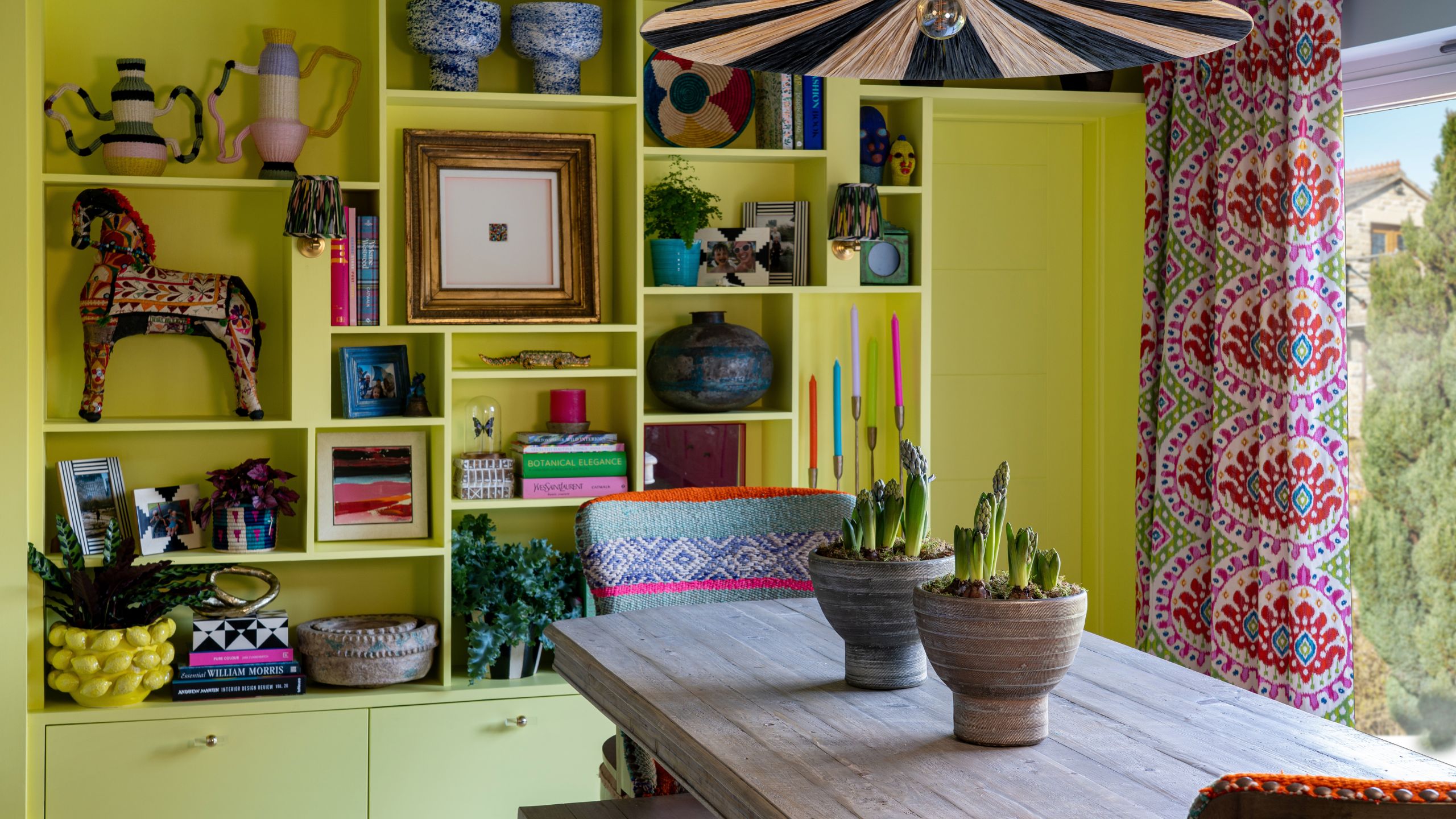 Vibrant dining room with chartreuse built-in shelves, a rustic wooden table with potted plants, and colorful ikat curtains