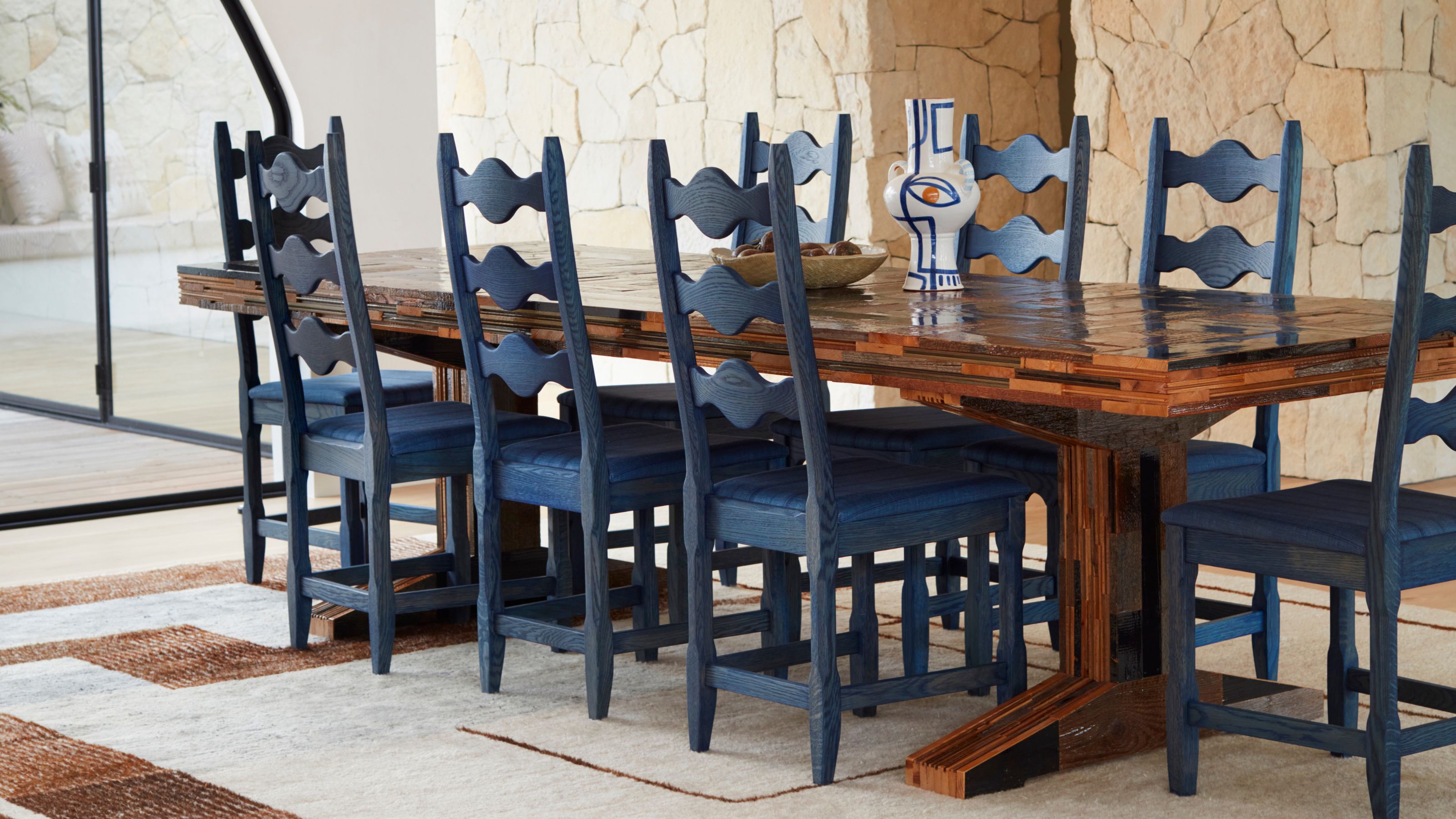 A dining space with stone wall on the right, white walls and a curved window. There is a large wood dining table with blue wooden chairs around and a chandelier above.