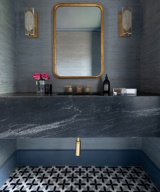 Half bathroom with blue textured wallpaper, gold sconces and mirror, marble effect vanity, patterned floor tiles,