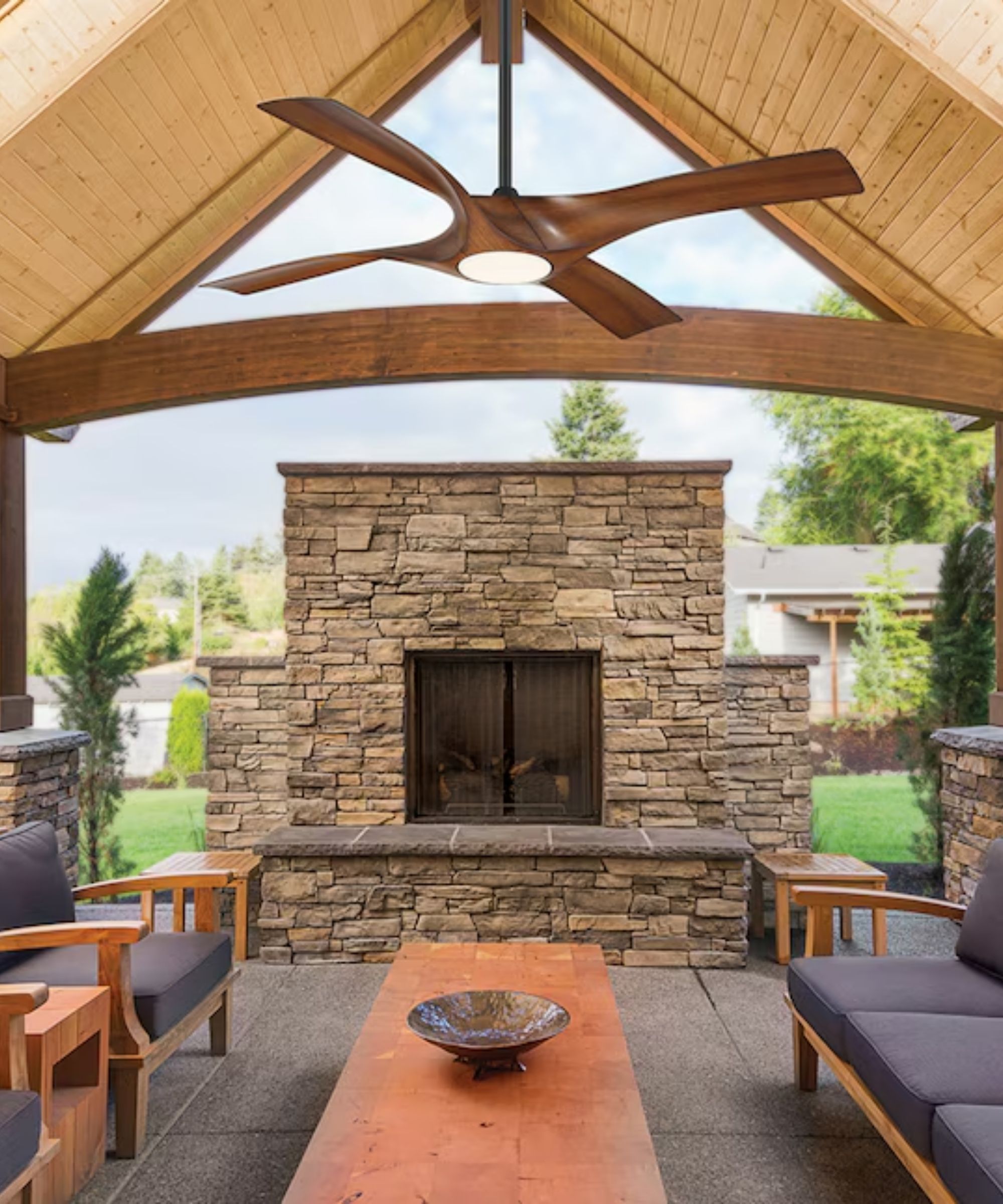 A Lowe's wooden ceiling fan hanging in an outdoor space, with a large, long table, sloped wooden ceilings, and a large stone fireplace, with wooden and navy seating to either side of the table.