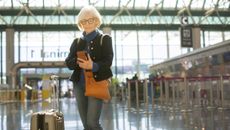 A senior woman stands at the airport with her luggage while checking her phone. 