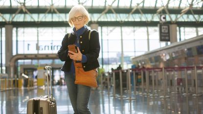 A senior woman stands at the airport with her luggage while checking her phone. 