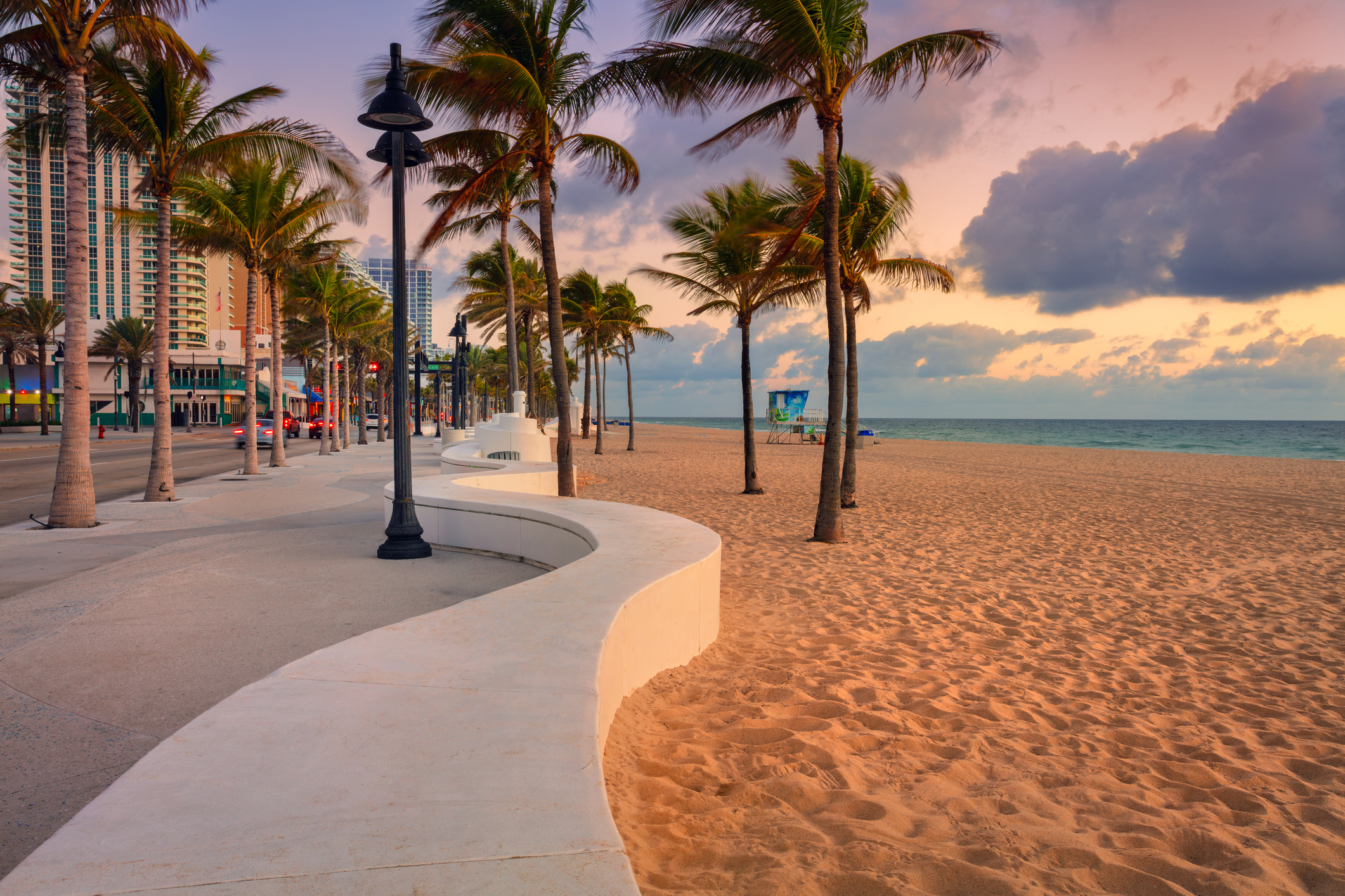 Palm trees border a sandy beach in Fort Lauderdale, Florida, with a curved walkway featuring white benches and street lamps. High-rise buildings line the opposite side.