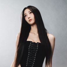 A portrait of singer-songwriter EJAE, wearing a lace-up corset and standing in front of a gray background.