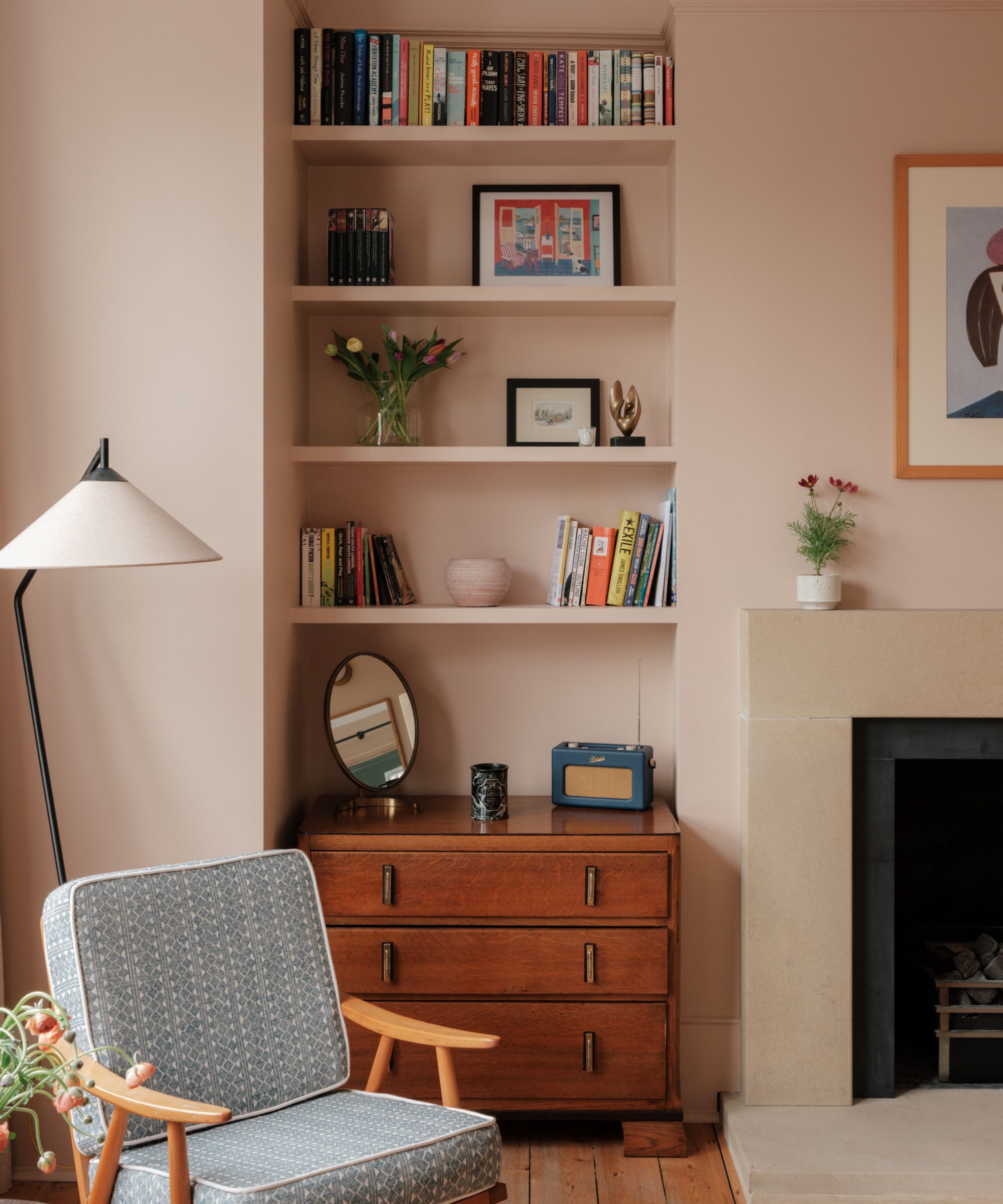 Living room with plaster pink walls, bookshelf nook with books and decor, a midcentury wooden chest of drawers with a radio, a midcentury lounge chair and floor lamp and a fireplace with an abstract print above