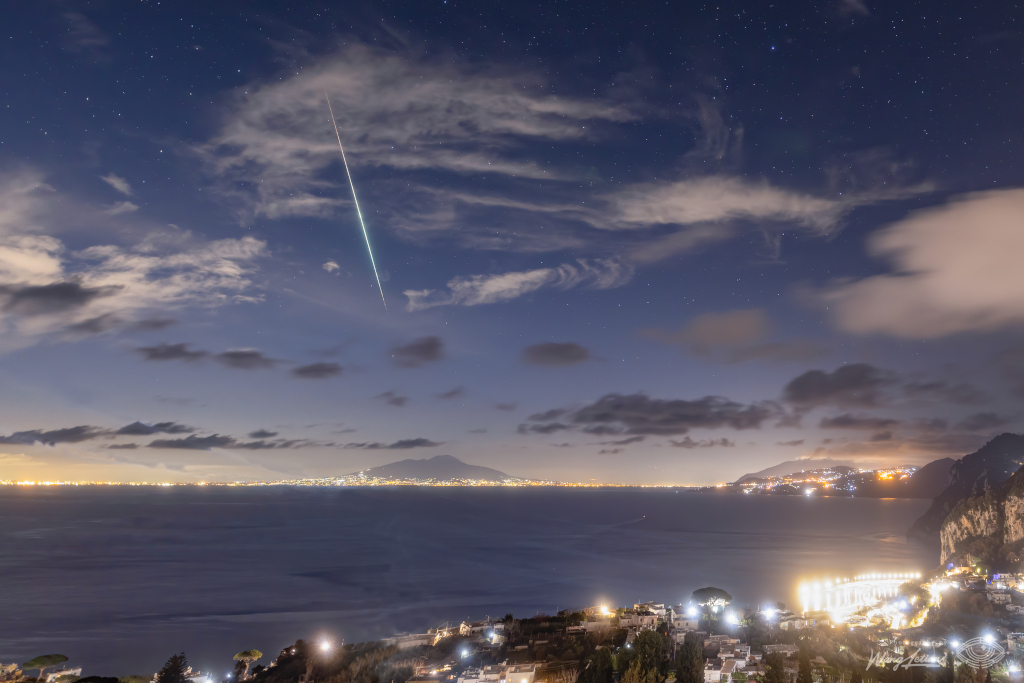 the hilly shoreline of an expansive bay is ilt with city lights next to the dark blue body of water. above, the sky fades from lit yellow to deep starry blue as clouds hang overhead. A meteor streaks downward in a trail of green.