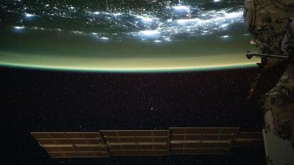 A large solar array is seen at the bottom of this image floating in space as the top half of the image shows Earth with a green sheen over its surface