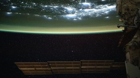 A large solar array is seen at the bottom of this image floating in space as the top half of the image shows Earth with a green sheen over its surface