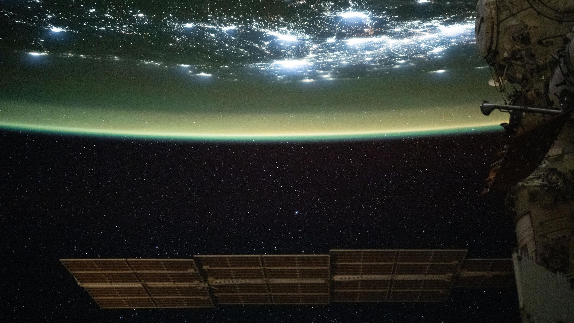 A large solar array is seen at the bottom of this image floating in space as the top half of the image shows Earth with a green sheen over its surface