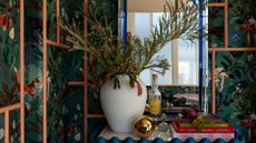 A close-up of a nook with tropical wallpaper and pastel peach framing, a blue side cabinet with books on top, and a white vase with feathered stems in front of a mirror