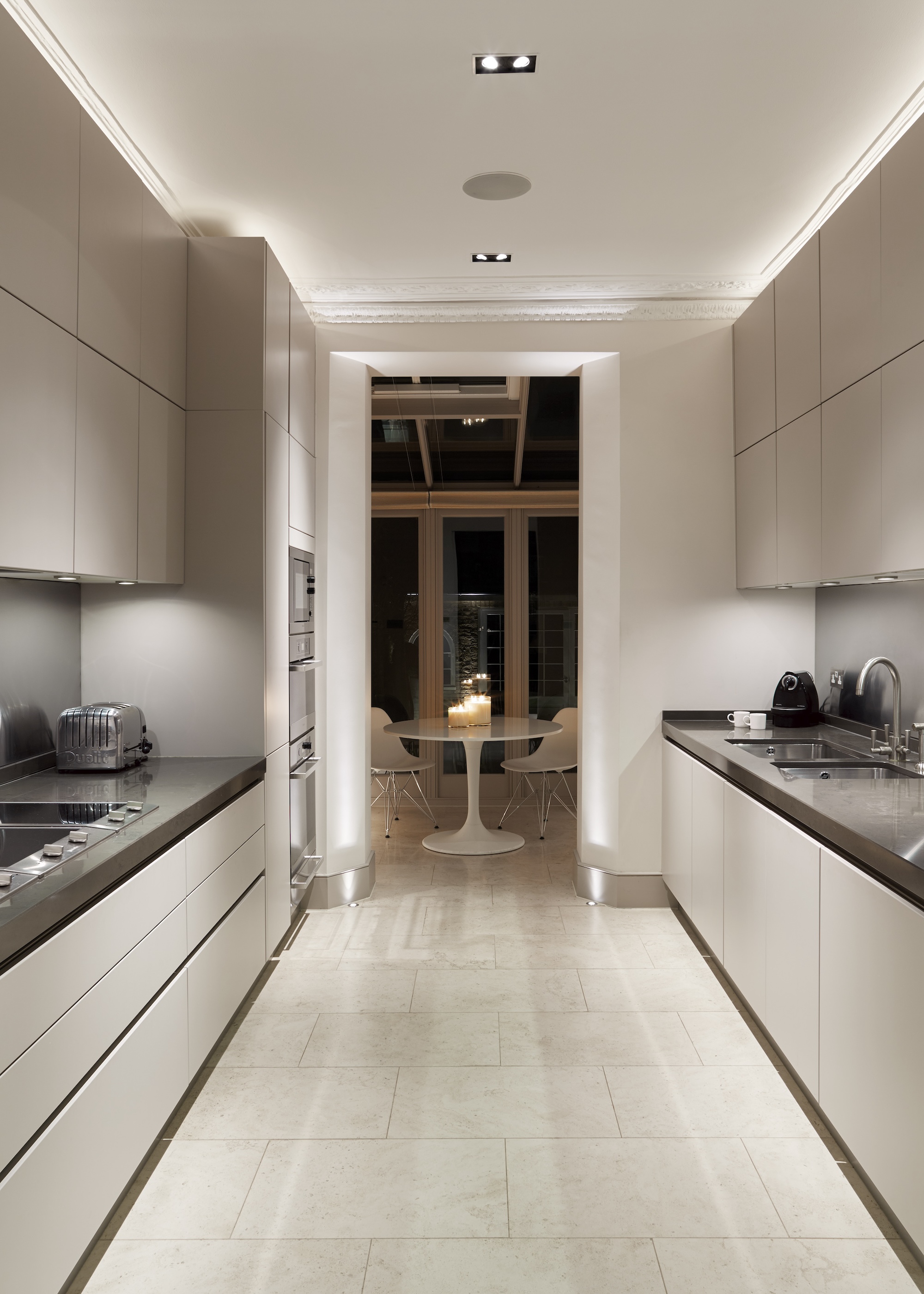 A long and narrow kitchen with LED lights above the cabinets. You can see a view of the countertops in the kitchen as well as a path that leads to the dining area.