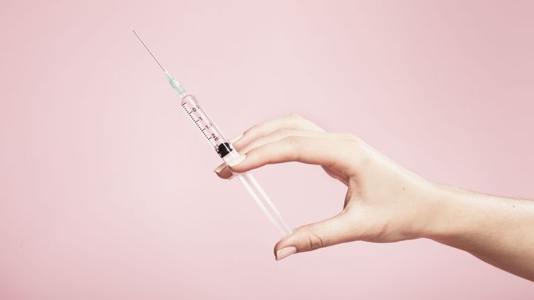 A hand holding a syringe set against a pink background.