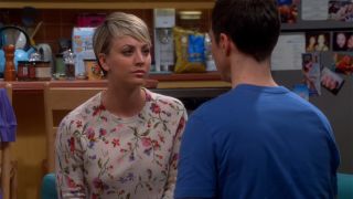 Kaley Cuoco sits on the couch and has a stare off with Jim Parsons as Penny and Sheldon in the big bang theory. She is in a floral shirt and has cut her hair short. He wears a tee.