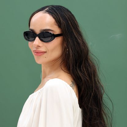 Zoë Kravitz standing in front of a step and repeat wall wearing a white saint laurent dress and sunglasses