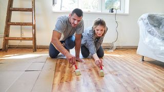 Two people sat on hardwood floors sealing it with paintbrushes.