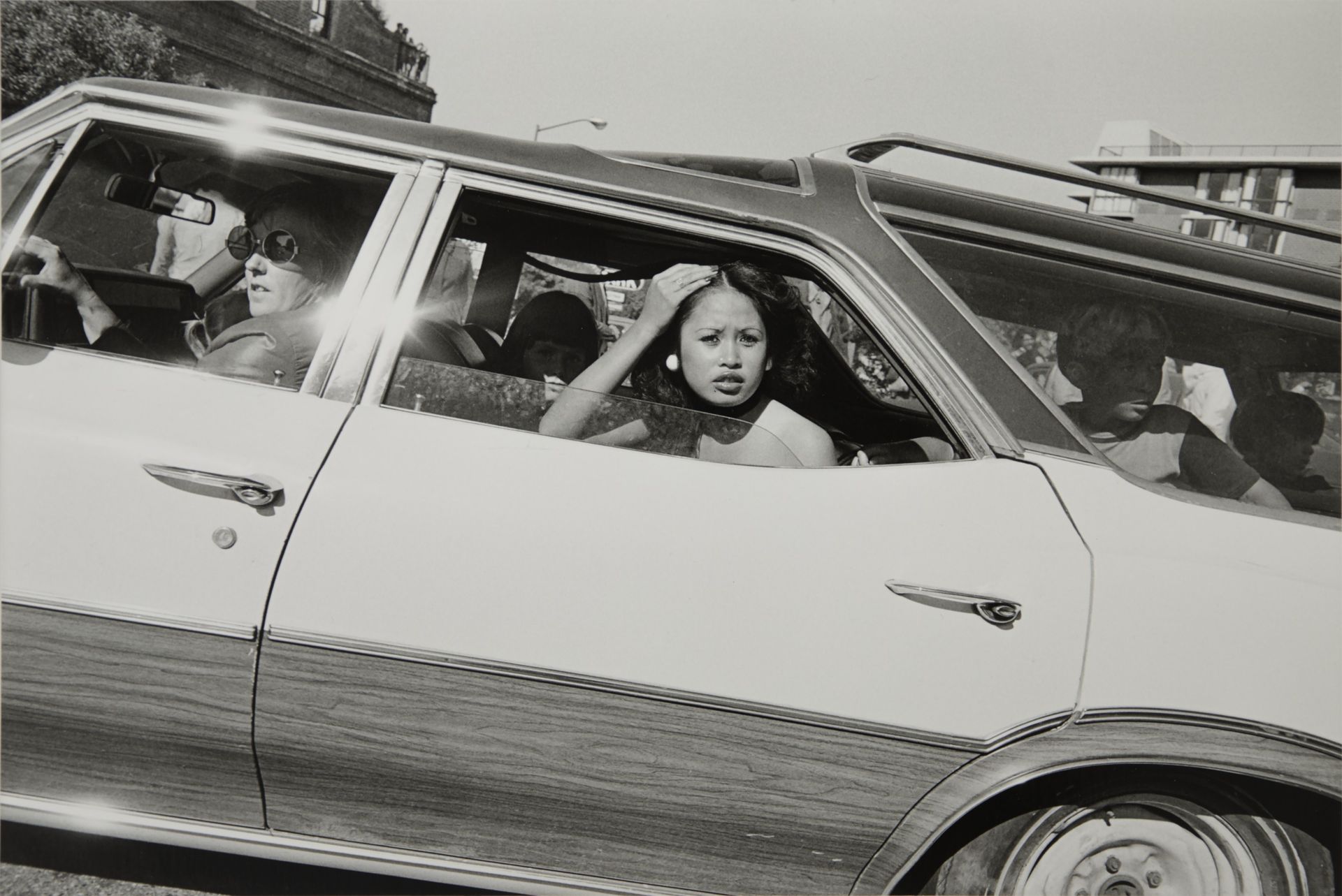 A black and white photograph taken through a car window of a young woman leaning out, her hand raised to her hair, as other passengers sit behind her.