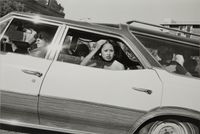 A black and white photograph taken through a car window of a young woman leaning out, her hand raised to her hair, as other passengers sit behind her.