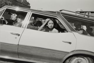 A black and white photograph taken through a car window of a young woman leaning out, her hand raised to her hair, as other passengers sit behind her.