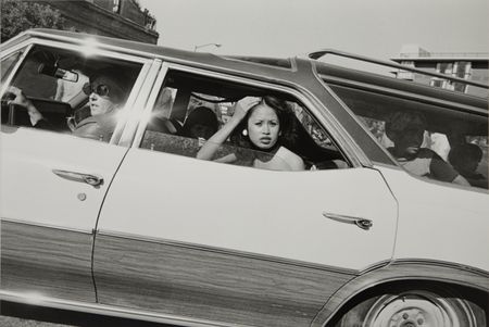 A black and white photograph taken through a car window of a young woman leaning out, her hand raised to her hair, as other passengers sit behind her.