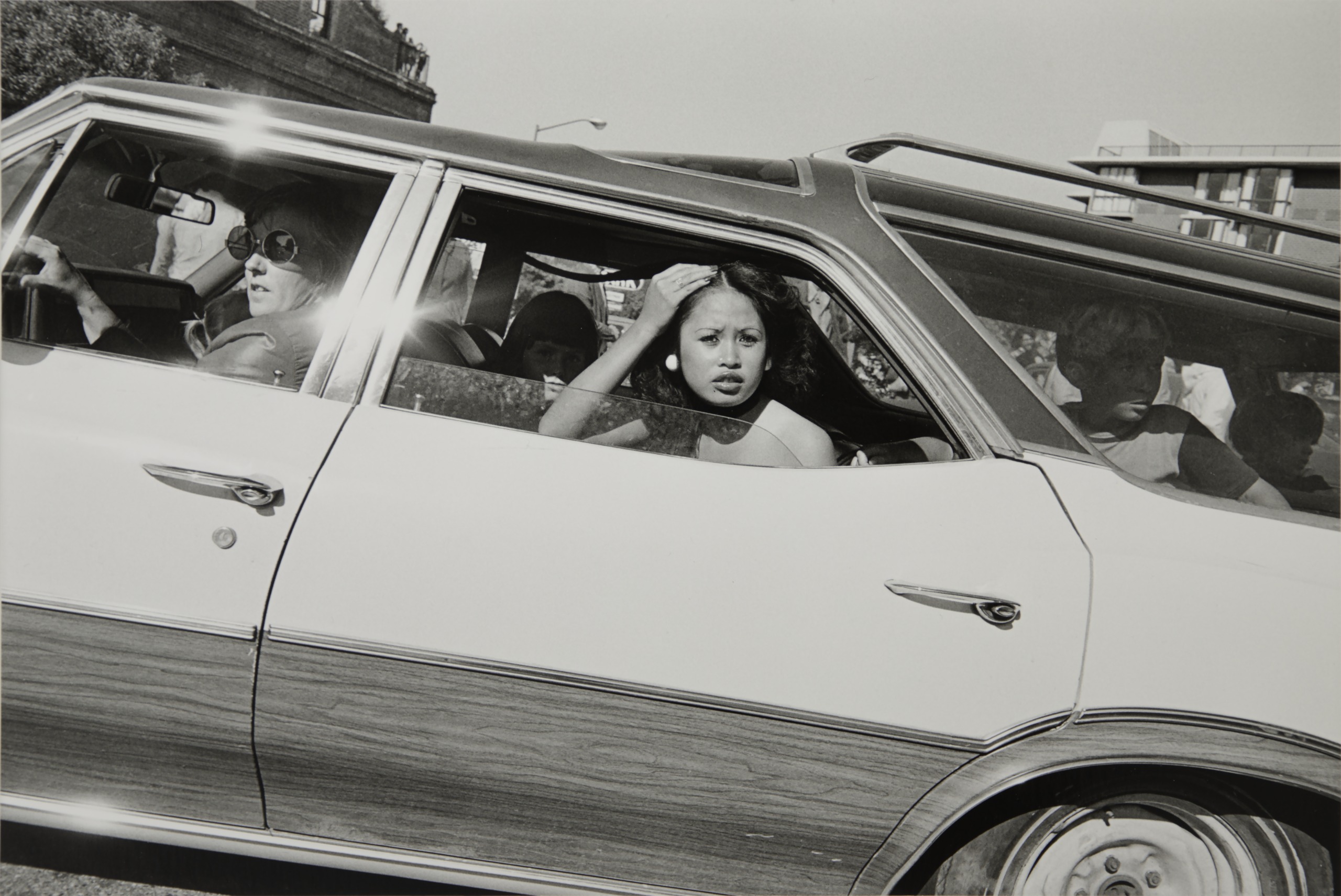 A black and white photograph taken through a car window of a young woman leaning out, her hand raised to her hair, as other passengers sit behind her.