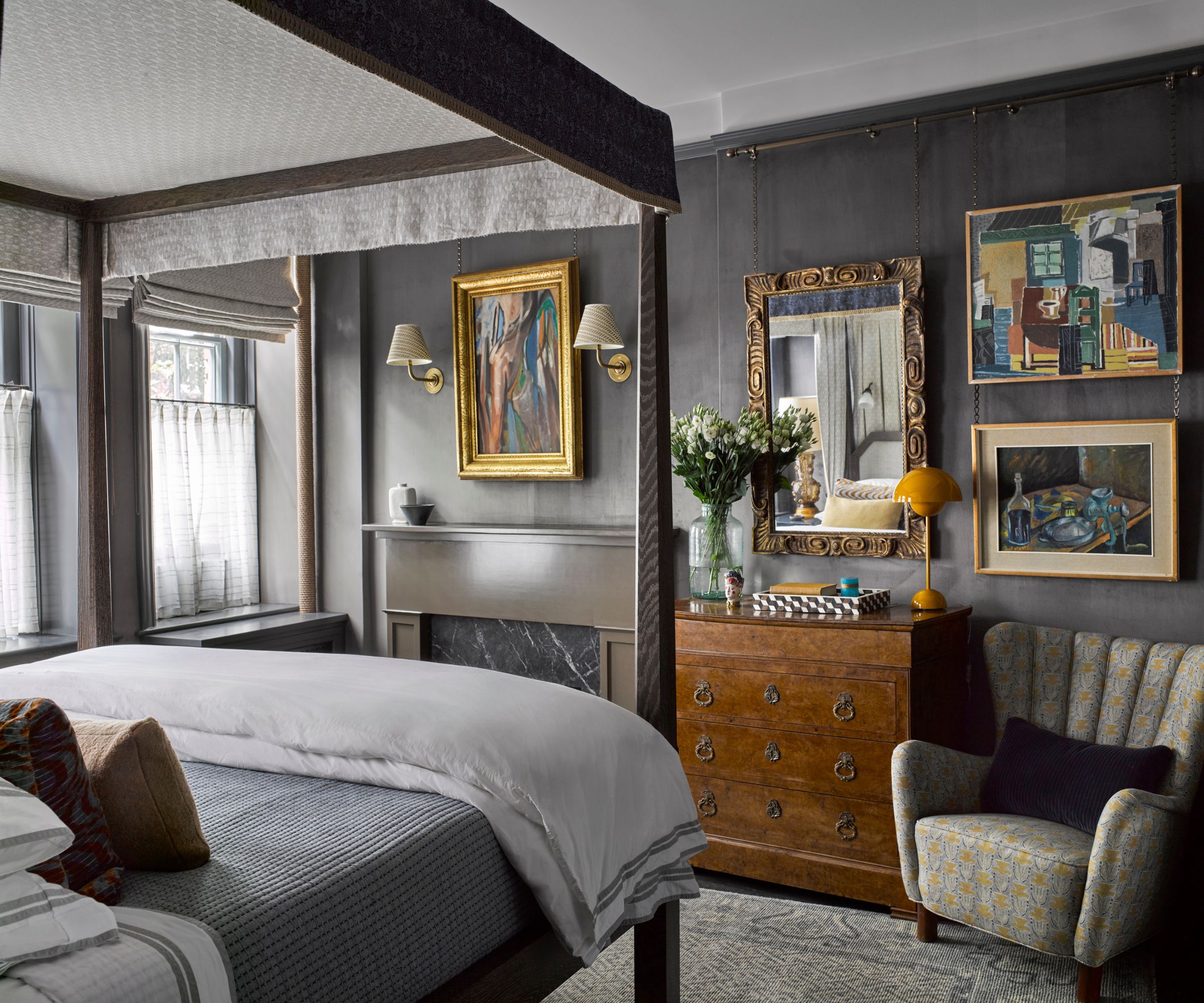A moody, bedroom with a dark wood four-poster bed, layered neutral bedding, vintage artwork, an antique dresser with a mirror, and a patterned armchair against gray walls