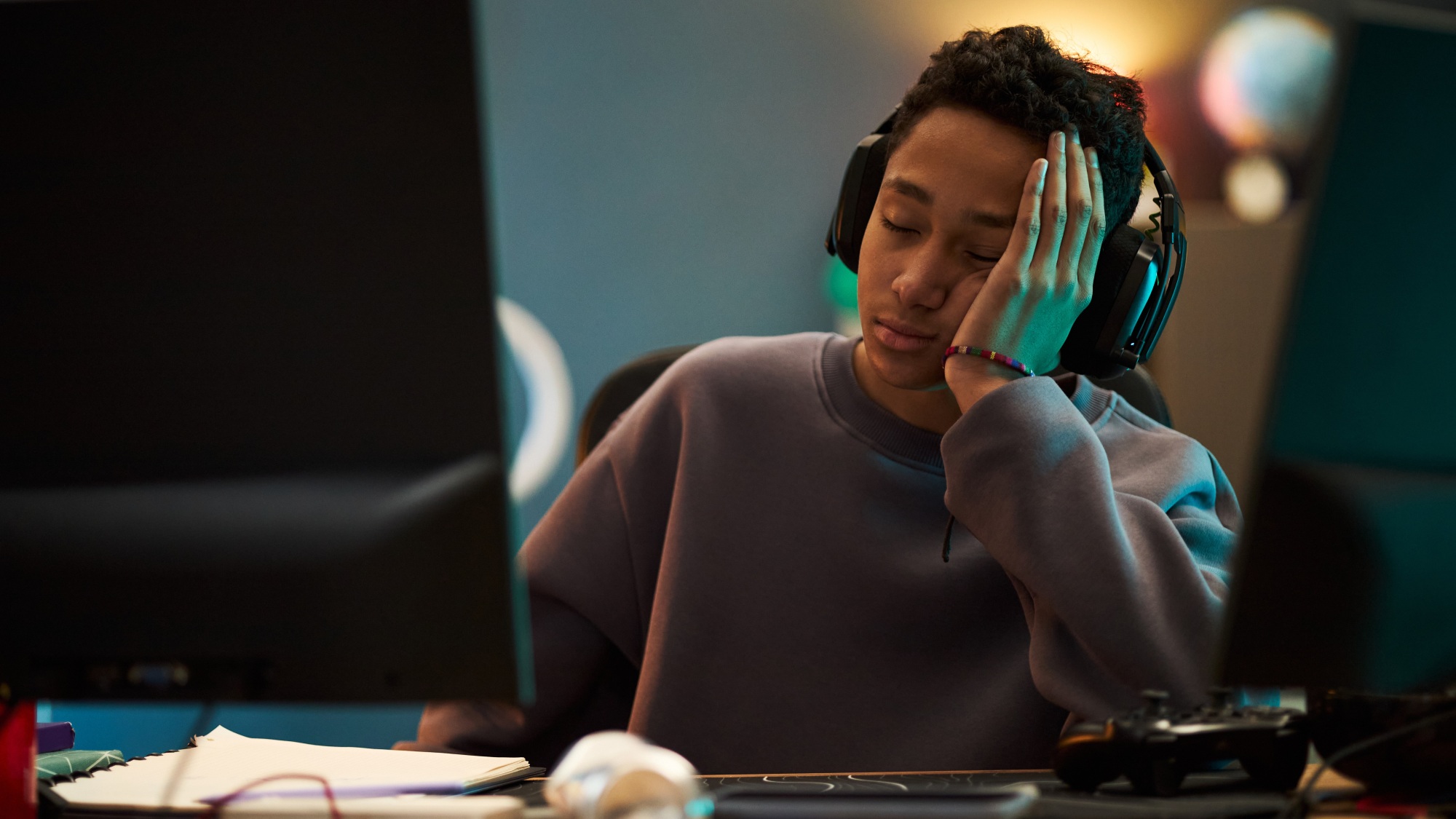 Teenager boy wearing headphones sitting at desk, resting head on hand appearing tired while gaming on computer monitor with game controller and papers visible on table