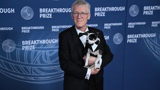 A man with gray hair wearing a black and white tuxedo holds a small black and white dog.