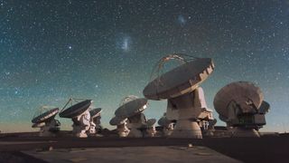 a dozen large round radio antenna dishes point upwards into a starry night sky