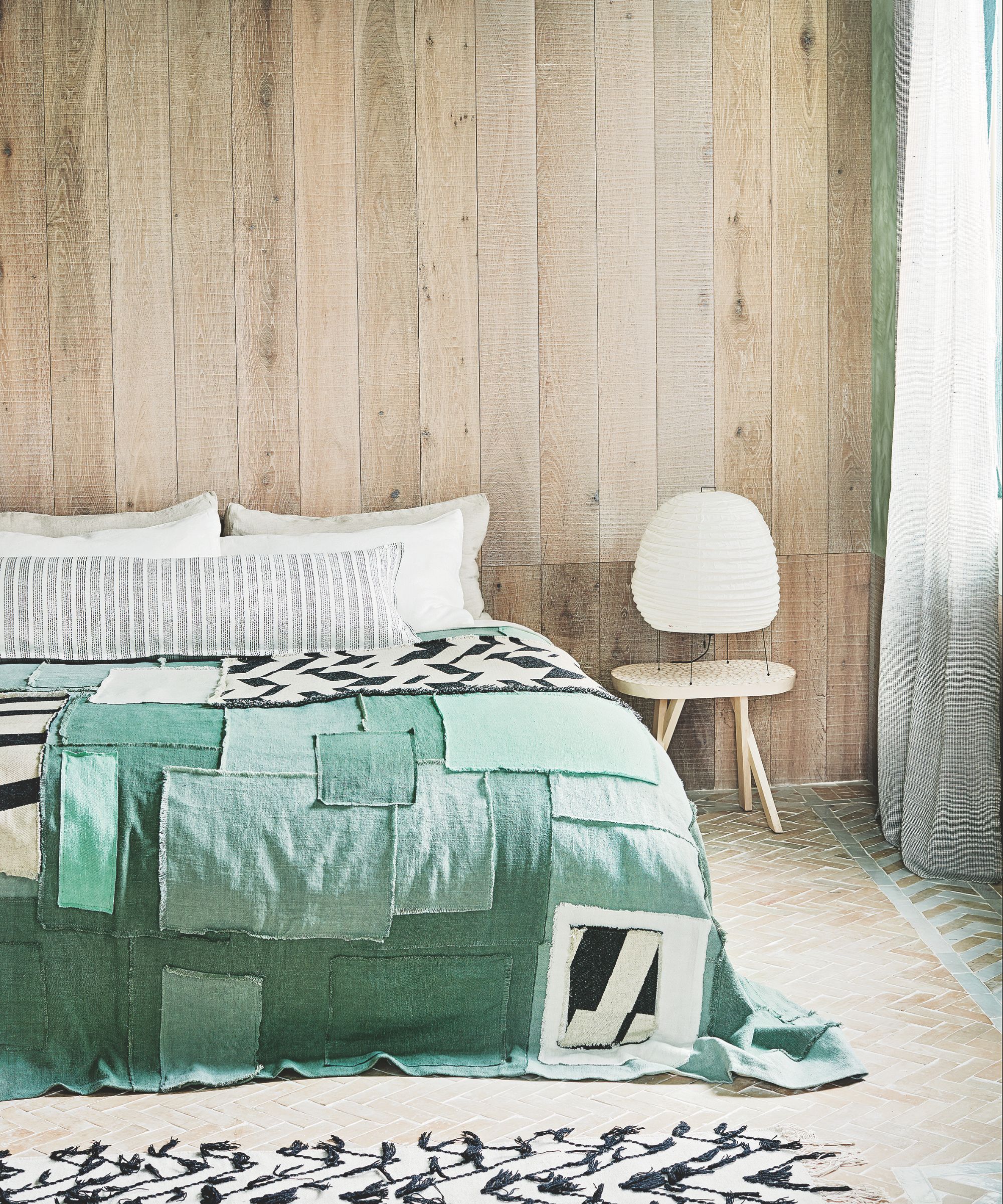 A light wood panelled bedroom with tiled flooring and a double bed with green patchwork quilt, white pillows, a white chair, and a white and black rug.