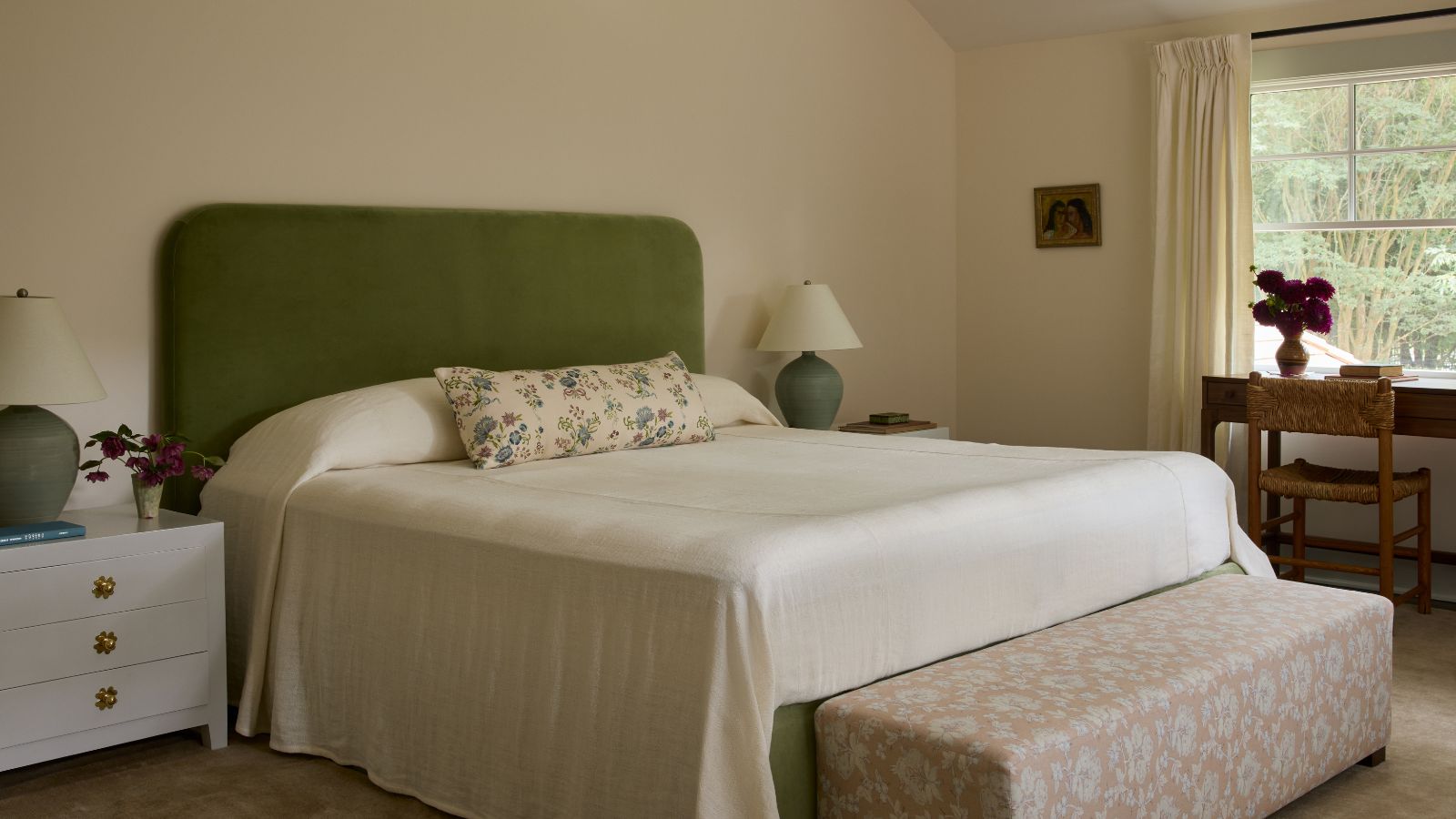 Primary bedroom with warm off-white walls, a green upholstered headboard, floral accent textiles, and natural wood furnishings, creating a soft and relaxed atmosphere.
